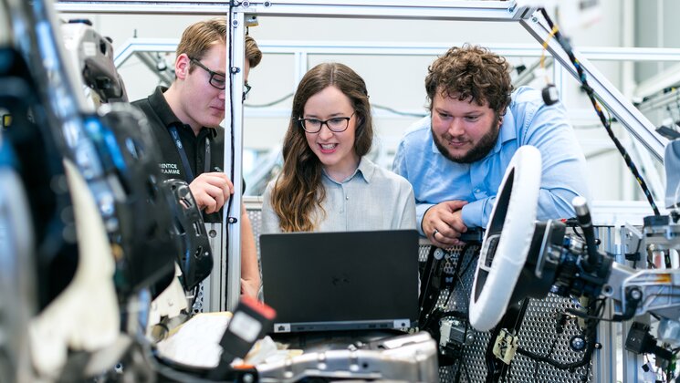 Three people looking at computer screen while standing near vehicle that they are building.